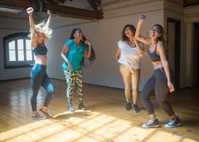 PARTNER GAIN COMPANY WORKOUT Four diverse women joyfully dancing together in a sunlit studio room. PARNERT WORKOUT WEDNESAY GAINS HUSTLE FITNESS BELFAST BEST GYM NUMBER 1 JOIN TODAY FREE FIRST COUPLE SESSION