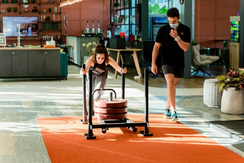 Woman pushing weights on a sled inside a modern gym with trainer observing.