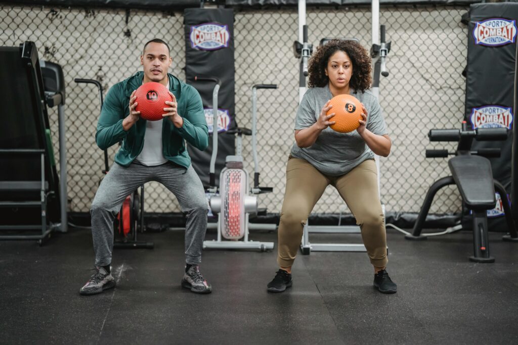 Full body sporty ethnic male coach and plus sized African American female in activewear squatting with fitness balls in hands in modern gym